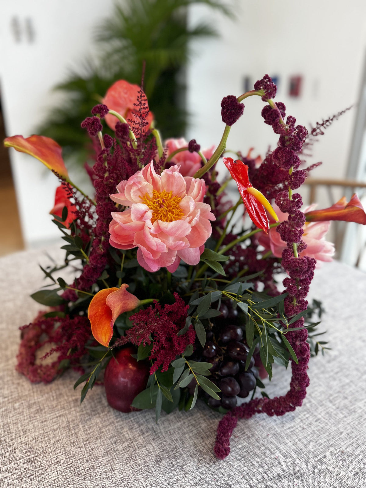 Coral peony, orange calla lilies, red anthurium and burgundy amaranth with grapes and an apple