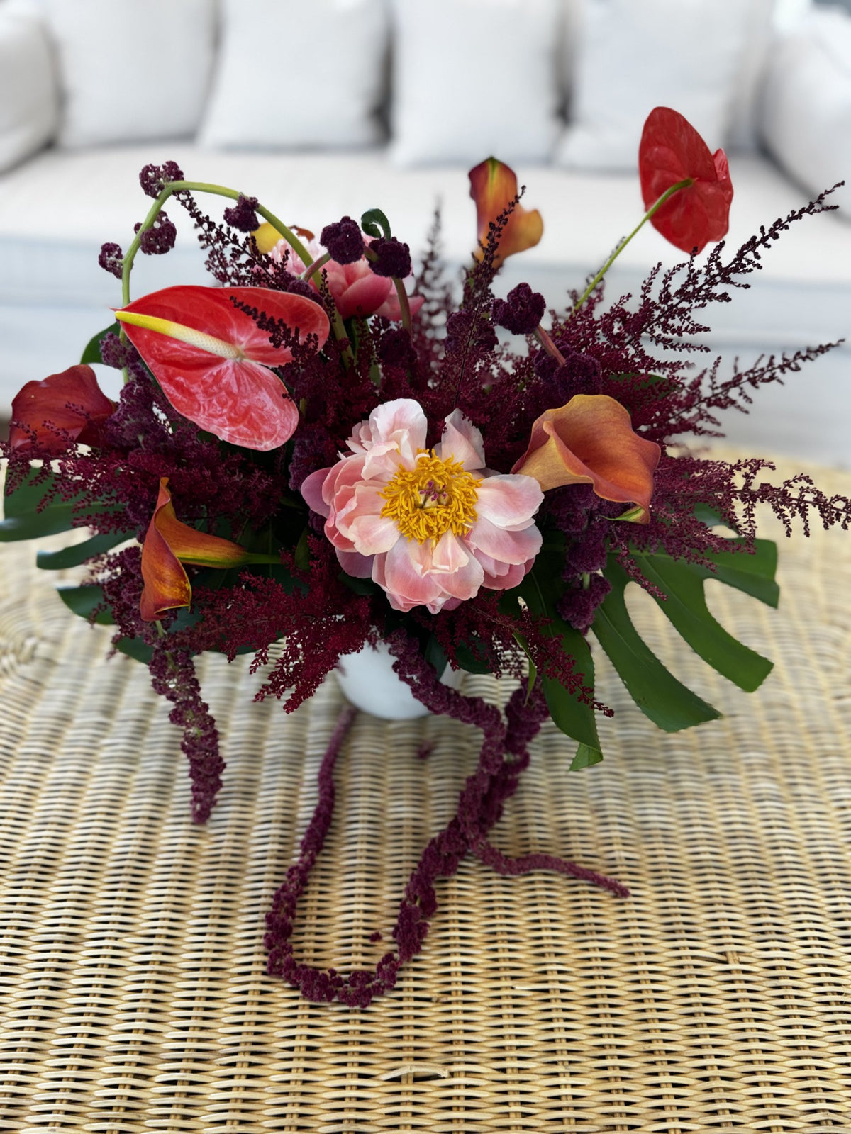 Coral peony, red anthurium, orange calla lilies and burgundy astilbe on a rattan table in a white room