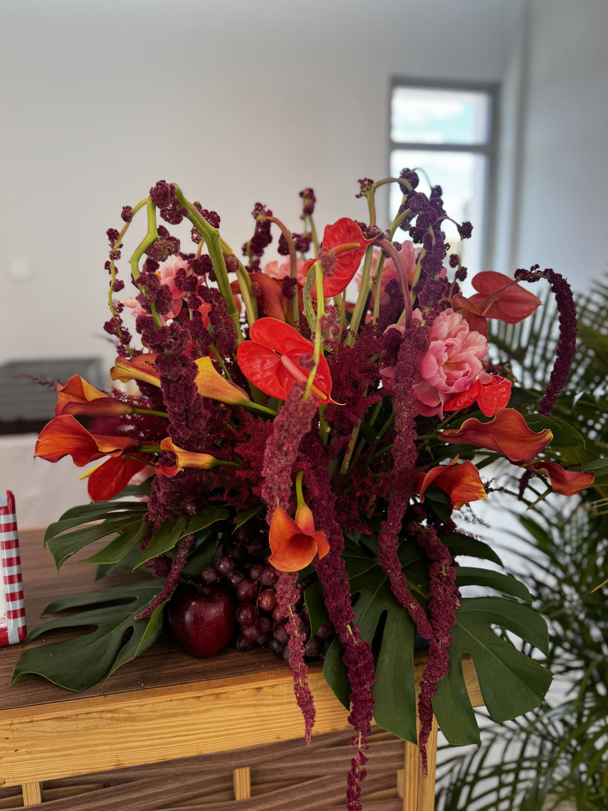 Tall arrangement of burgundy amaranth, red anthurium, orange calla lilies and monstera with grapes and apples