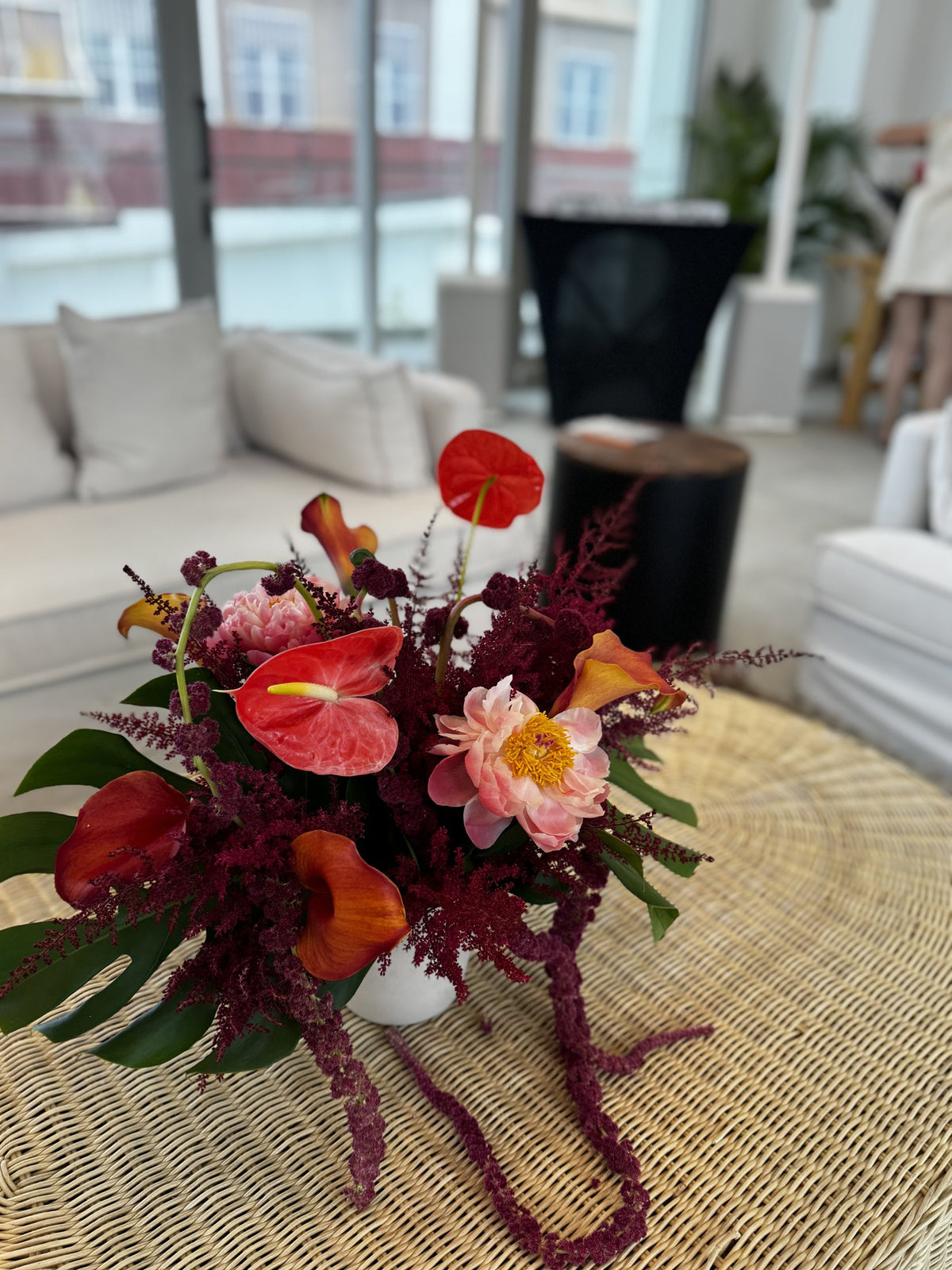 Coral peony, red anthurium and burgundy amaranth in a white vase on a rattan table in a sunny living room