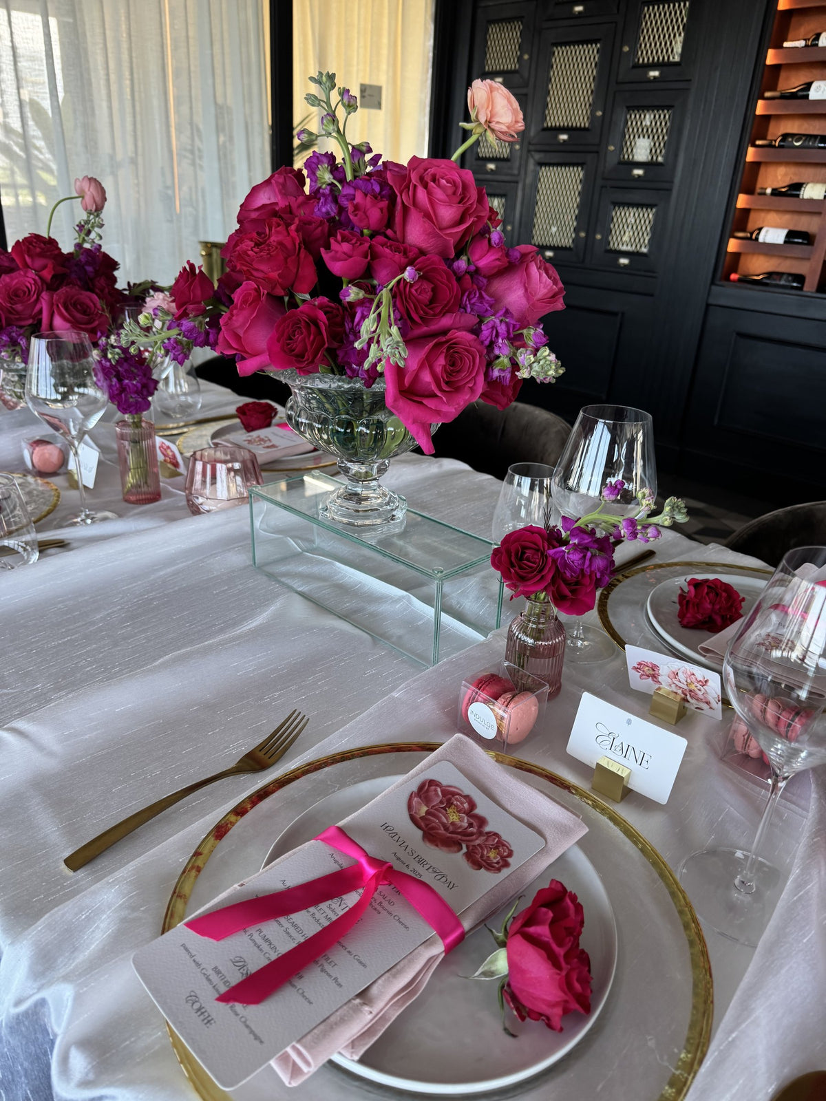 Fuchsia roses and purple stock in a crystal pedestal vase over a set table