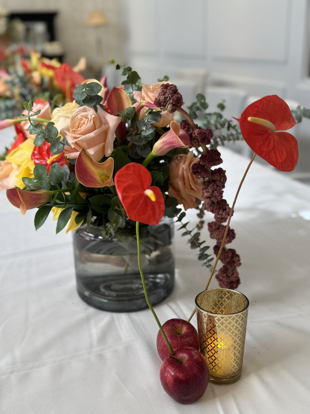 Red anthurium, peach roses and hanging amaranth in a smoked glass vase with apples