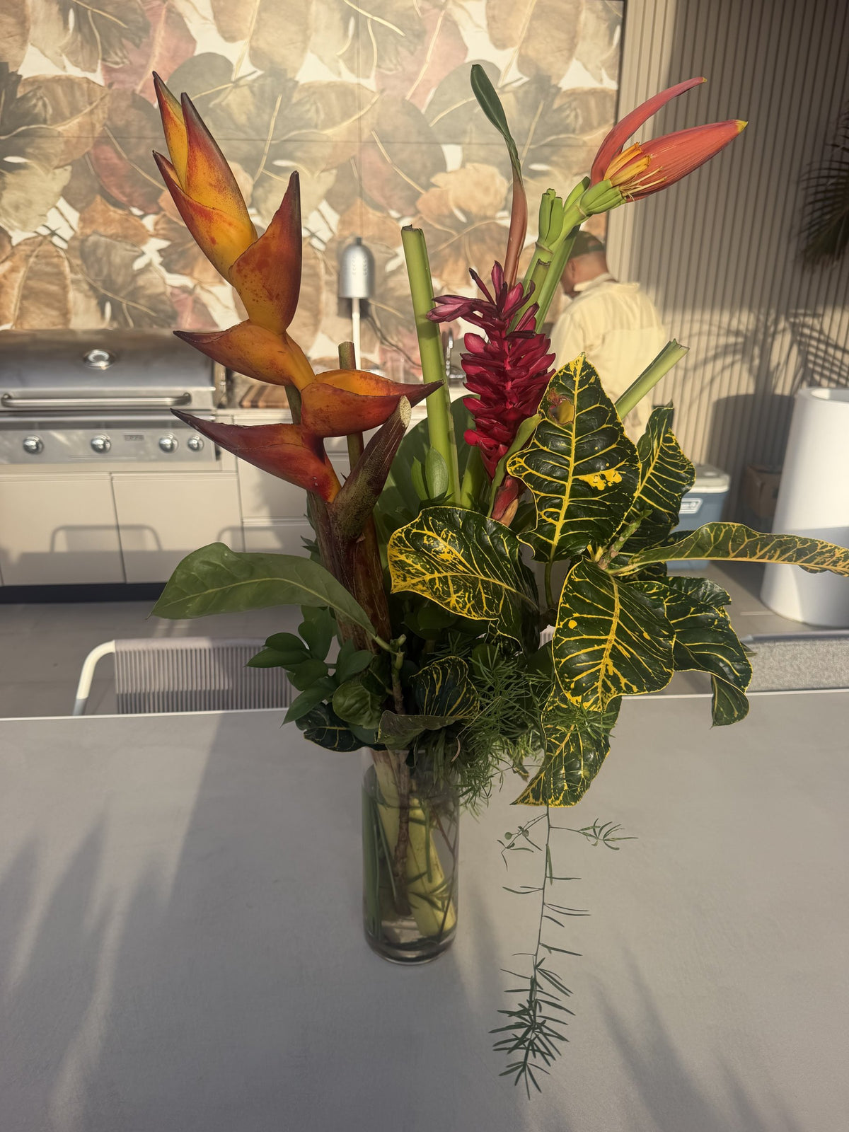 Heliconia, red ginger and croton leaves in a tall glass vase on an outdoor counter
