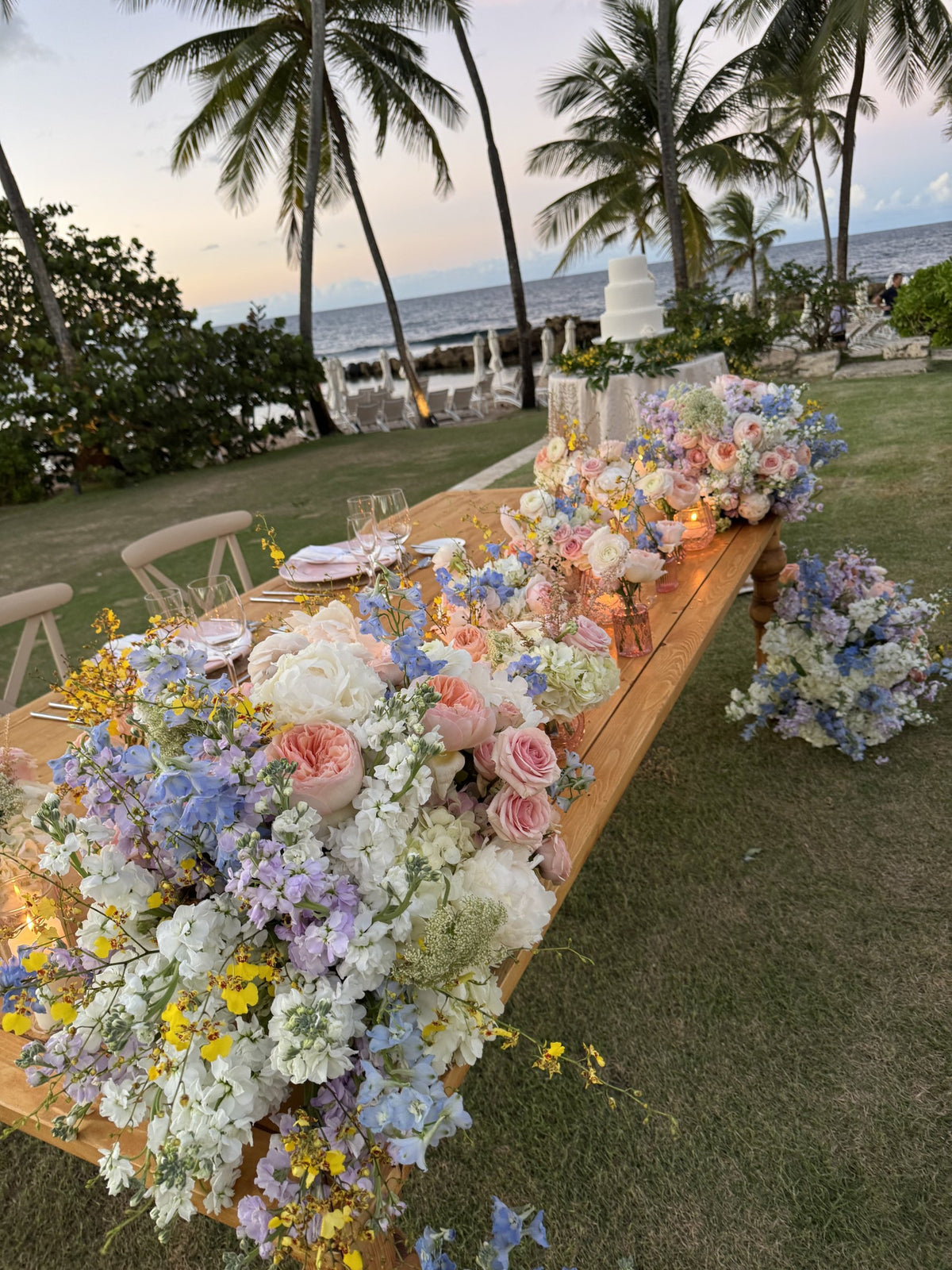 Long wooden table covered in pastel roses, delphinium and orchids, palm trees behind