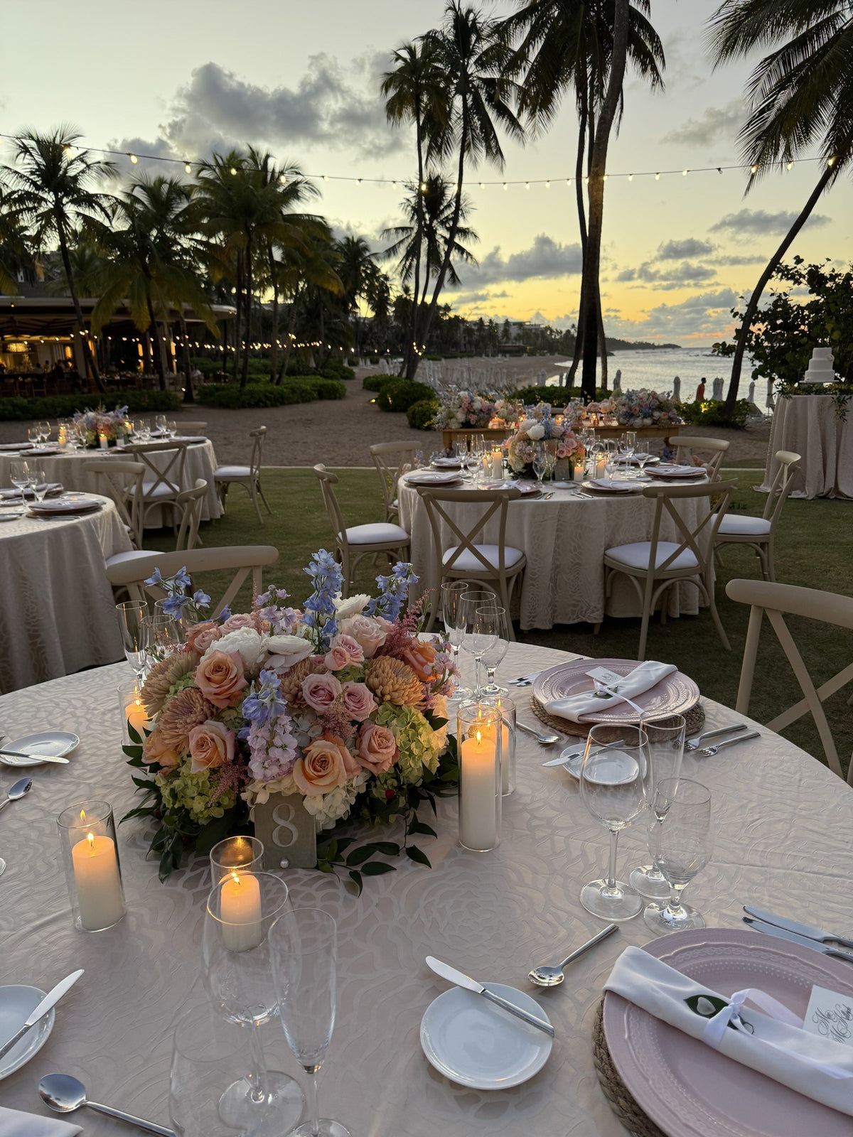 Round reception table with a pastel rose centerpiece and candles, palms and sea at dusk