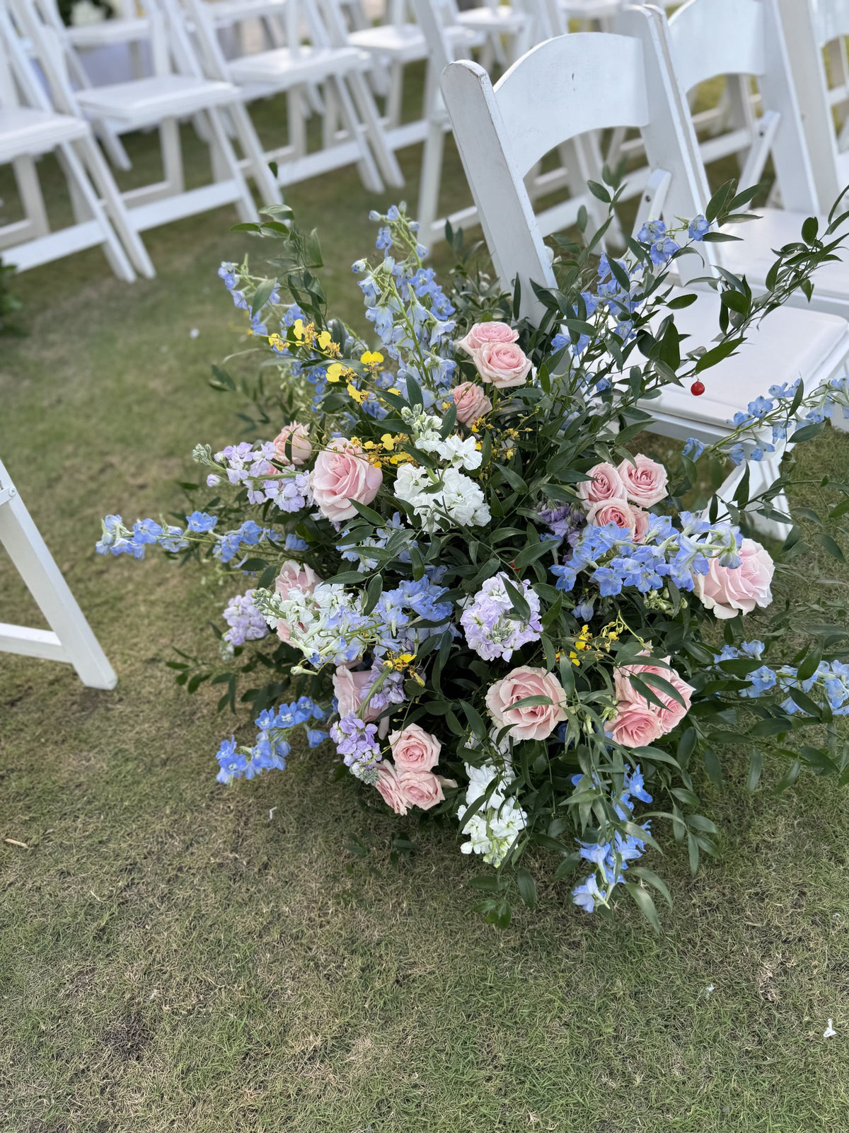 Ground arrangement of pastel roses and blue delphinium on the lawn by the ceremony chairs