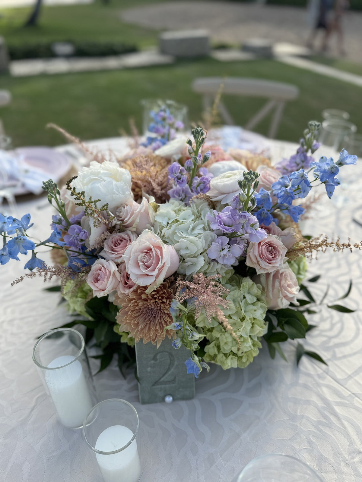 Low centerpiece of roses, hydrangea and delphinium with candles on a white tablecloth