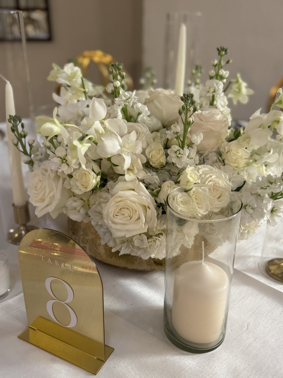 White roses, stock and hydrangea in a gold vessel with a gold table number