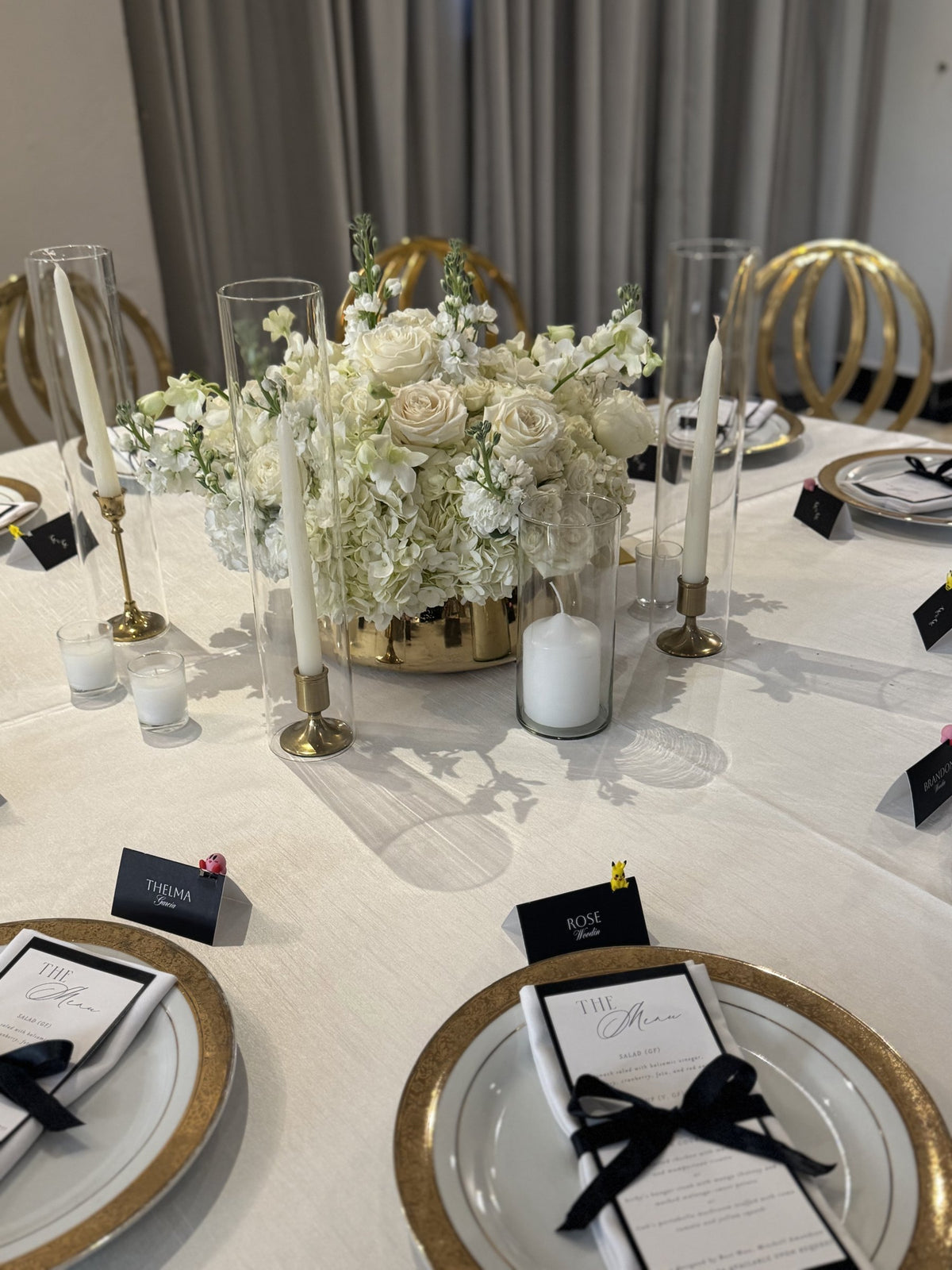 White roses and hydrangea in a gold vessel with taper candles and black-ribbon menus