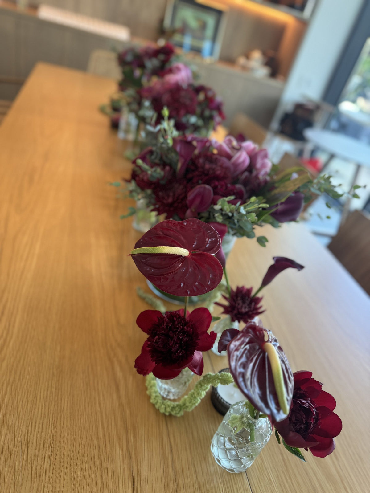 Long dining table with a row of burgundy arrangements and bud vases