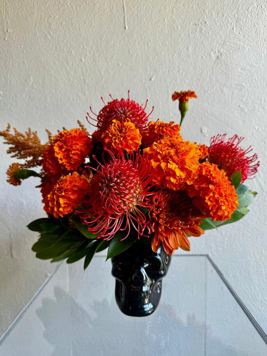 Halloween arrangement in a black skull with orange marigolds and red pincushion protea
