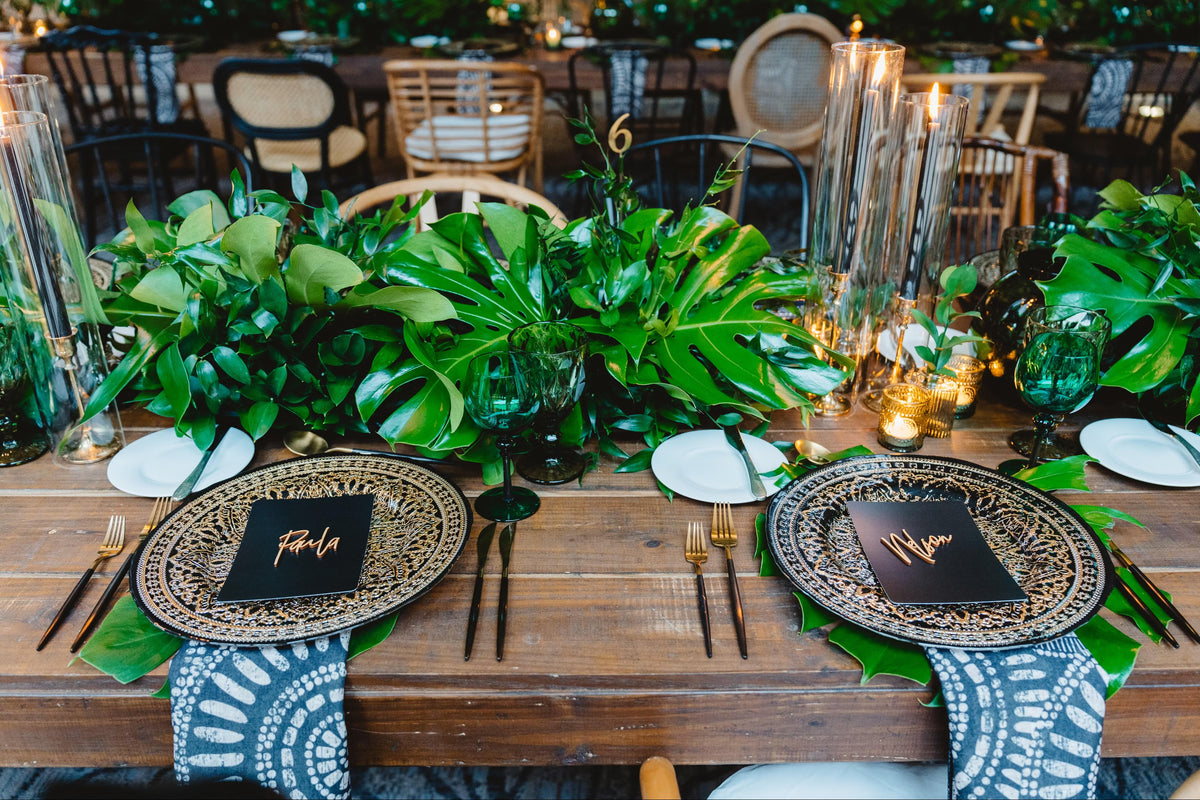 Monstera and palm table runner on dark wood, with tall candles and gold cutlery